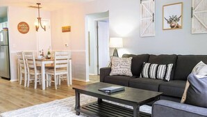 A living room with a dark brown sofa, a white rug, and a wooden coffee table. There is a dining area with a white table and chairs. - Cliffside Cottage 3 - Updated - Near Silver Dollar (Branson)