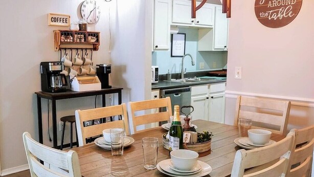 A dining area with a wooden table set for four, surrounded by white chairs. A coffee station with a coffee maker and mugs is visible in the background. - Cliffside Cottage 3 - Updated - Near Silver Dollar (Branson)