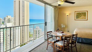 A hotel room with a balcony, a bed, a table with chairs, a ceiling fan, and a view of the city and ocean.