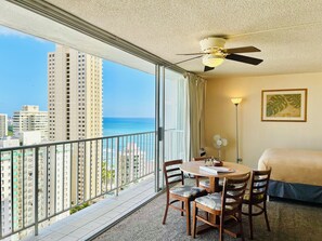 A hotel room with a balcony, a bed, a table with chairs, a ceiling fan, and a view of the city and ocean. - "STEPS AWAY FROM THE WAIKIKI BEACH STUDIO WITH OCEAN VIEW" (Honolulu)