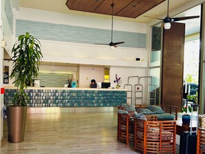 Un espace d’accueil comprenant un comptoir de réception avec un réceptionniste, une grande plante en pot et un coin salon avec des chaises en bois et une table.