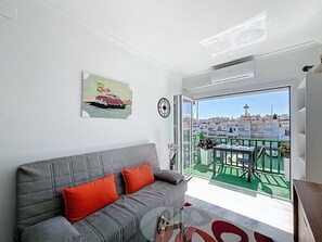Ein modernes Wohnzimmer mit einem grauen Sofa, roten Kissen, einem Glastisch und Blick auf eine Stadtlandschaft vom Balkon aus.