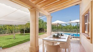 A covered patio with a table and chairs, a pool in the background, and a view of a well-maintained lawn.