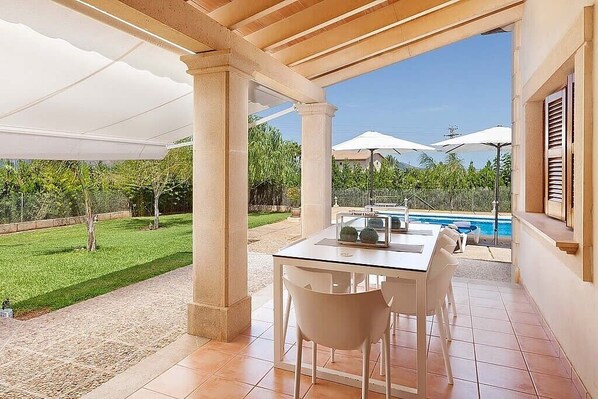 A covered patio with a table and chairs, a pool in the background, and a view of a well-maintained lawn.