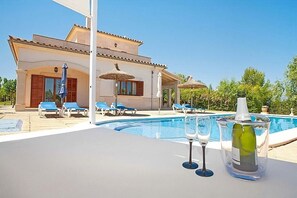 A poolside area with lounge chairs, a table with a bottle of wine and glasses, and a building with a tiled roof.