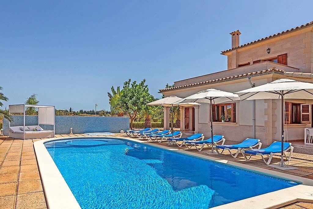 Une piscine avec des chaises longues et des parasols, un bâtiment doté d’un toit en tuiles et un ciel dégagé.