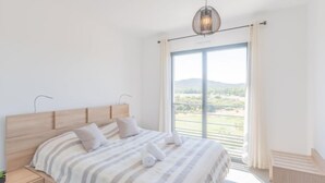 Ein Schlafzimmer mit einem großen Bett, einem Nachttisch, einer Hängeleuchte und Blick auf eine Landschaft durch eine Schiebetür.