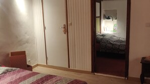 Een slaapkamer met een houten vloer, een bed met een beddeken met een patroon, een kledingkast en een klein tafeltje met boeken.