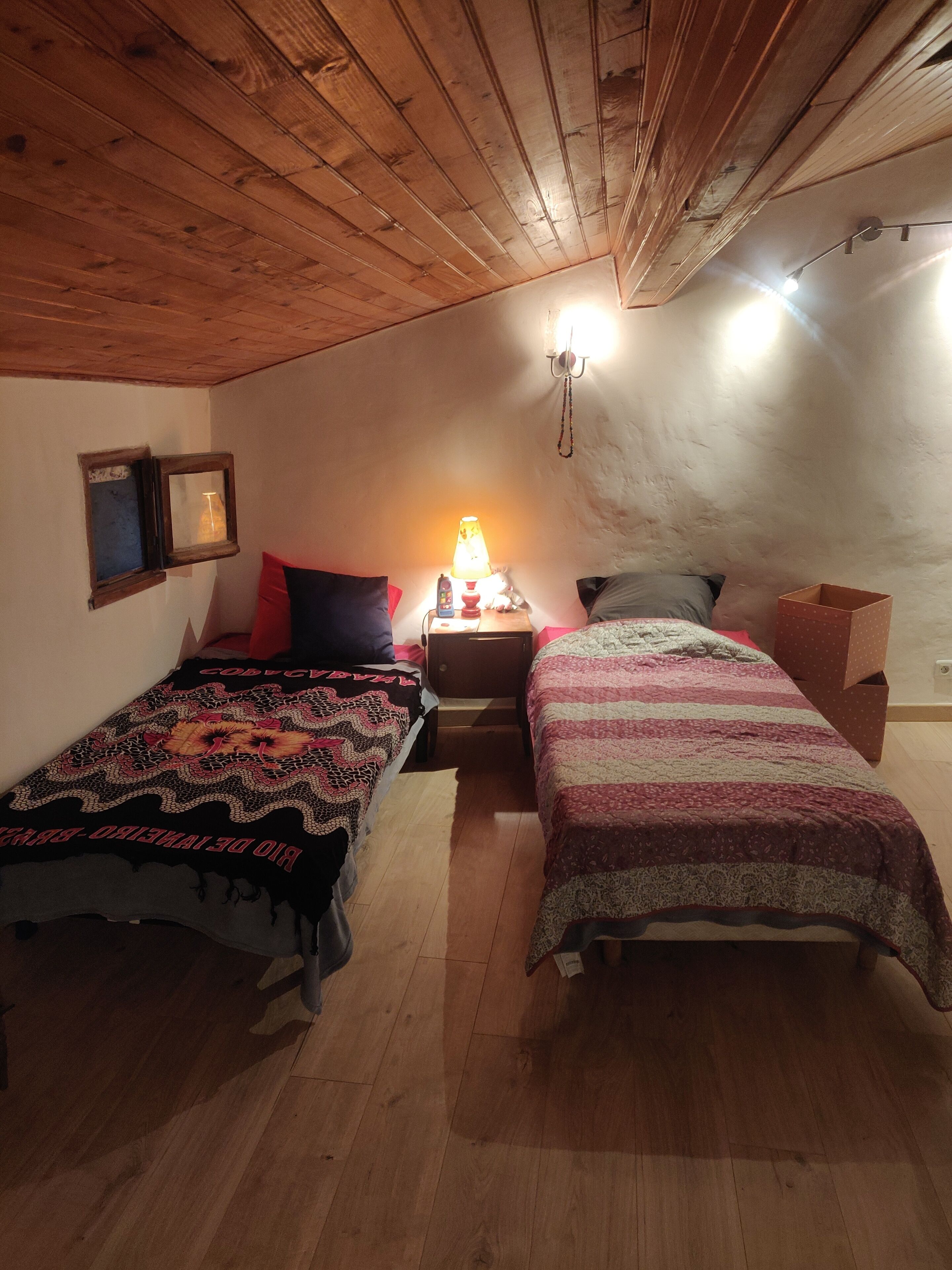 A cozy attic bedroom with two beds, wooden ceiling beams, a nightstand with a lamp, and a small window.