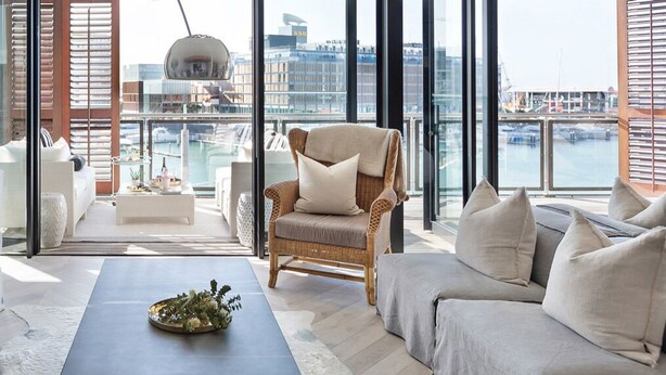 Un moderno salón con vista a un puerto deportivo, que incluye un conjunto de sofás, una mesa de centro y un sillón de mimbre.