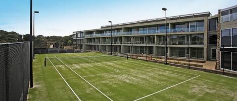 A tennis court adjacent to a modern building with large windows and a flat roof.