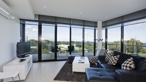 A modern living room with a large TV, black leather sofa, and a view of the city through floor-to-ceiling windows. - Whitewater Apartment 101 (TORQUAY)
