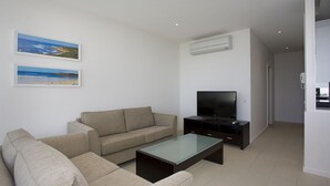 Un salon moderne avec un canapé beige, une table basse en verre et un téléviseur à écran plat posé sur un meuble foncé. Deux tableaux représentant des plages, encadrés, ornent le mur.