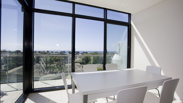 Une chambre moderne dotée d’une grande fenêtre en verre, de meubles blancs et offrant une vue sur le paysage urbain.