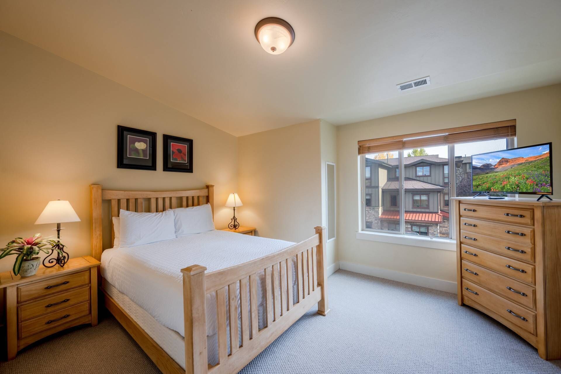 A bedroom with a bed, bedside tables, a dresser, a TV, and a window with a view of buildings.