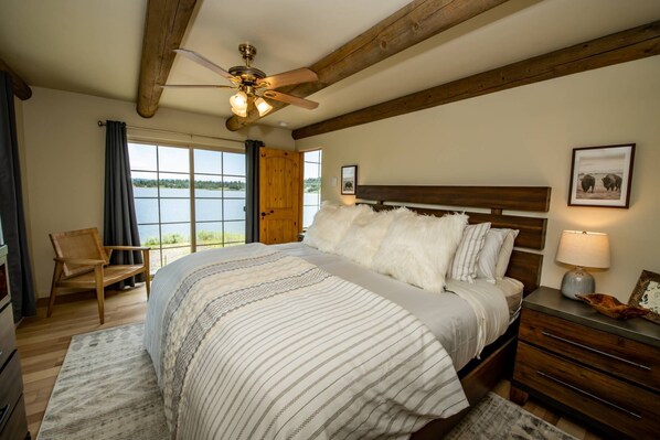 A bedroom with a large bed, wooden headboard, a ceiling fan, a window with a view of a lake, a chair, and a nightstand with a lamp.