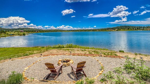 A lakeside view with a circular seating area featuring Adirondack chairs and a fire pit.