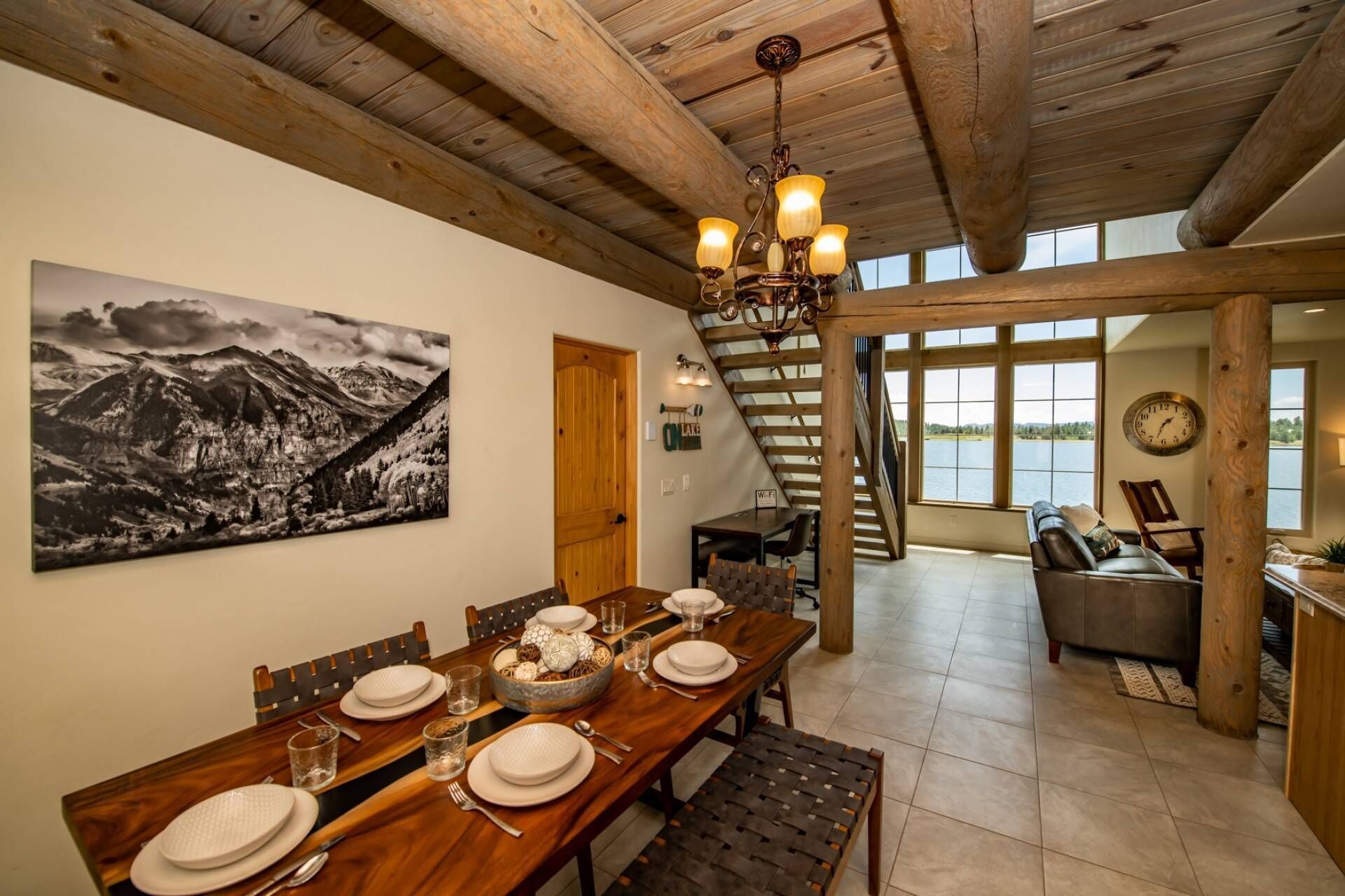 A dining area with a wooden table set for four, a chandelier, and a mountain landscape painting on the wall.