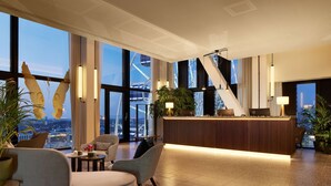 A modern hotel lobby with a reception desk, seating area, large windows, and a view of the cityscape.
