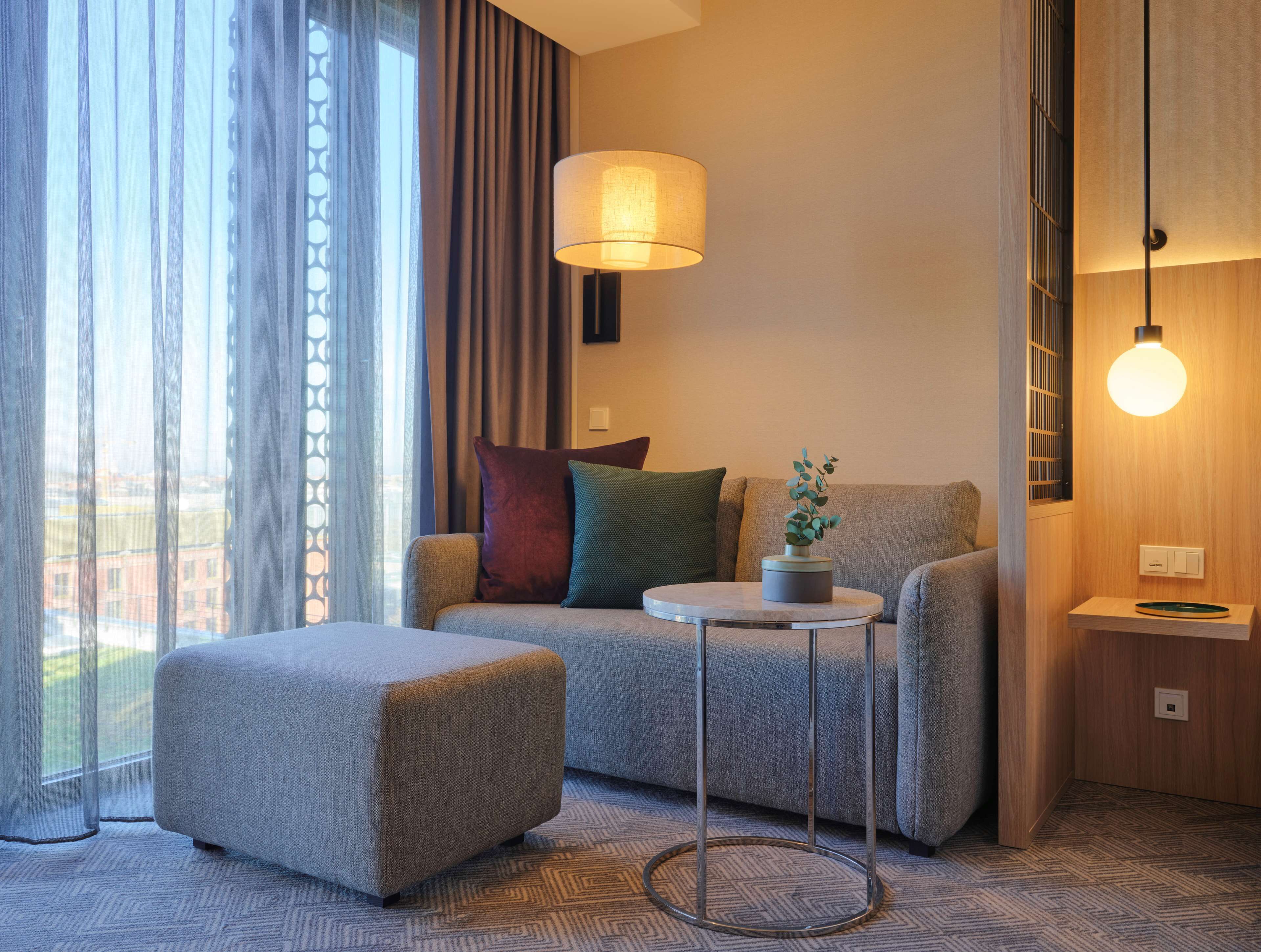 Quarto de hotel moderno com sofá cinzento, um banquinho a combinar, uma mesinha redonda com um vaso e uma janela ampla com cortinas leves.