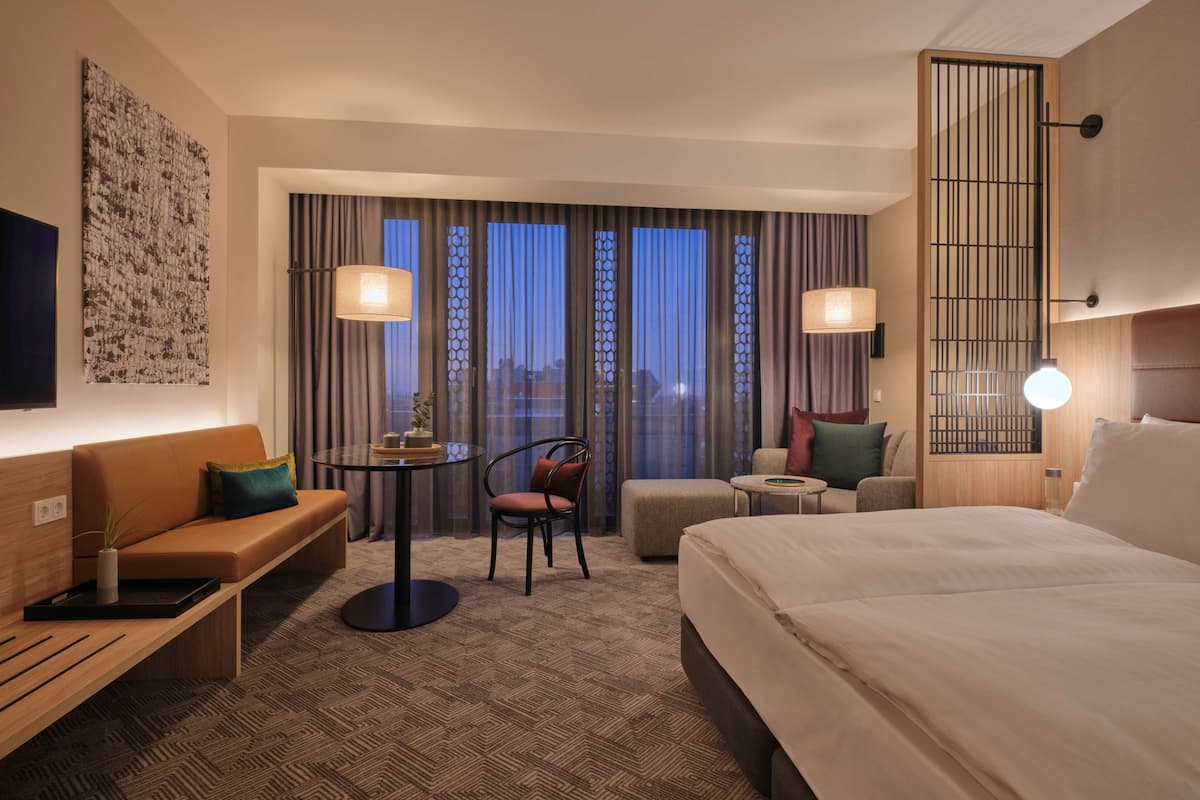 Ein Hotelzimmer mit einem großen Bett, einem Sofa, einem kleinen Tisch und einem Sessel. Vorhänge und ein Blick auf die Stadtlandschaft durch die Fenster.