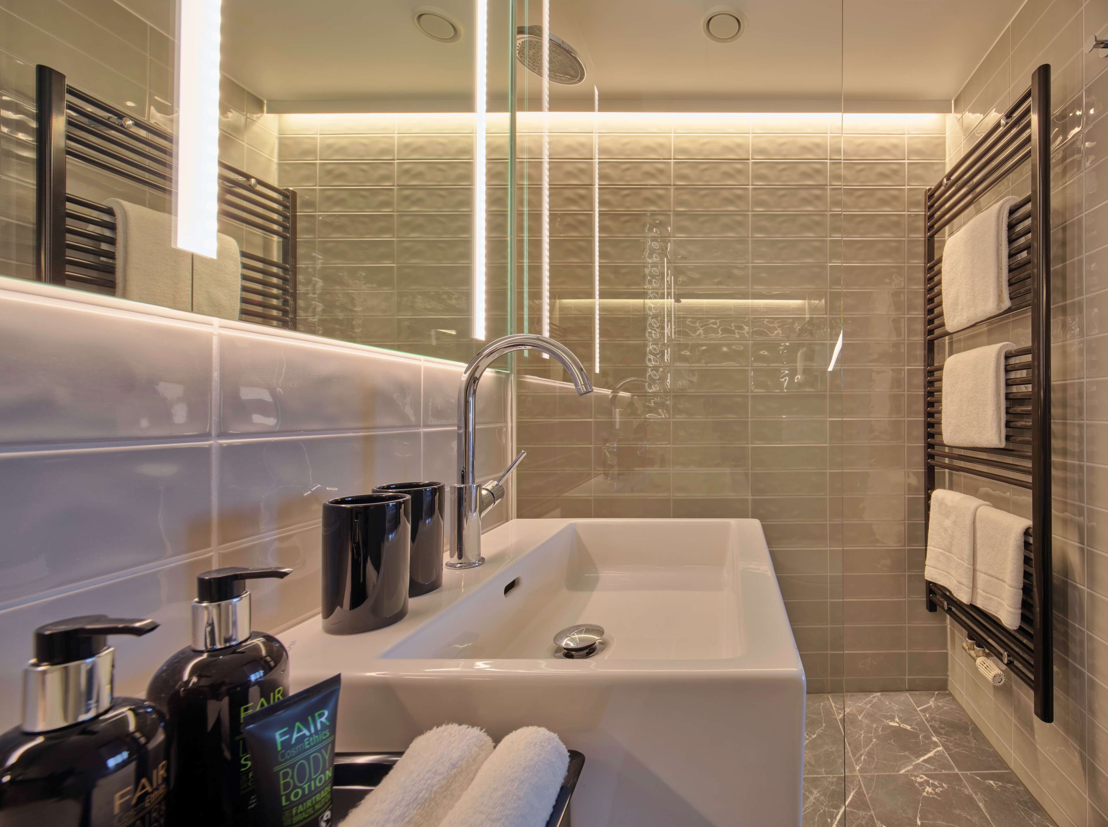 Ein modernes Badezimmer mit einer großen Badewanne, einer Handbrause und einer Duschkabine aus Glas. Auf dem Waschbecken stehen zwei schwarze Flaschen, und Handtücher hängen ordentlich auf einem Ablagegestell.