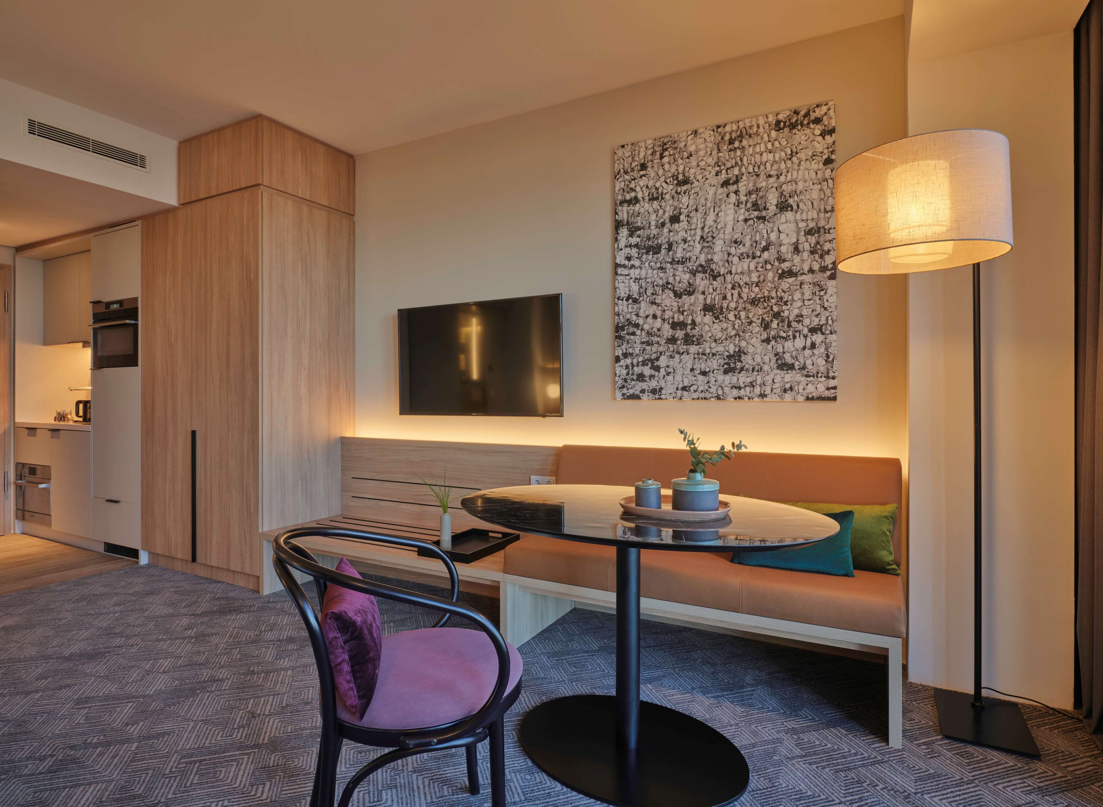 A modern hotel room with a dining area, a sofa, a TV, and a floor lamp.