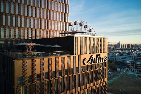 A high-rise building with a prominent 'Adina' sign, featuring a modern architectural design with vertical lines and a rooftop seating area.