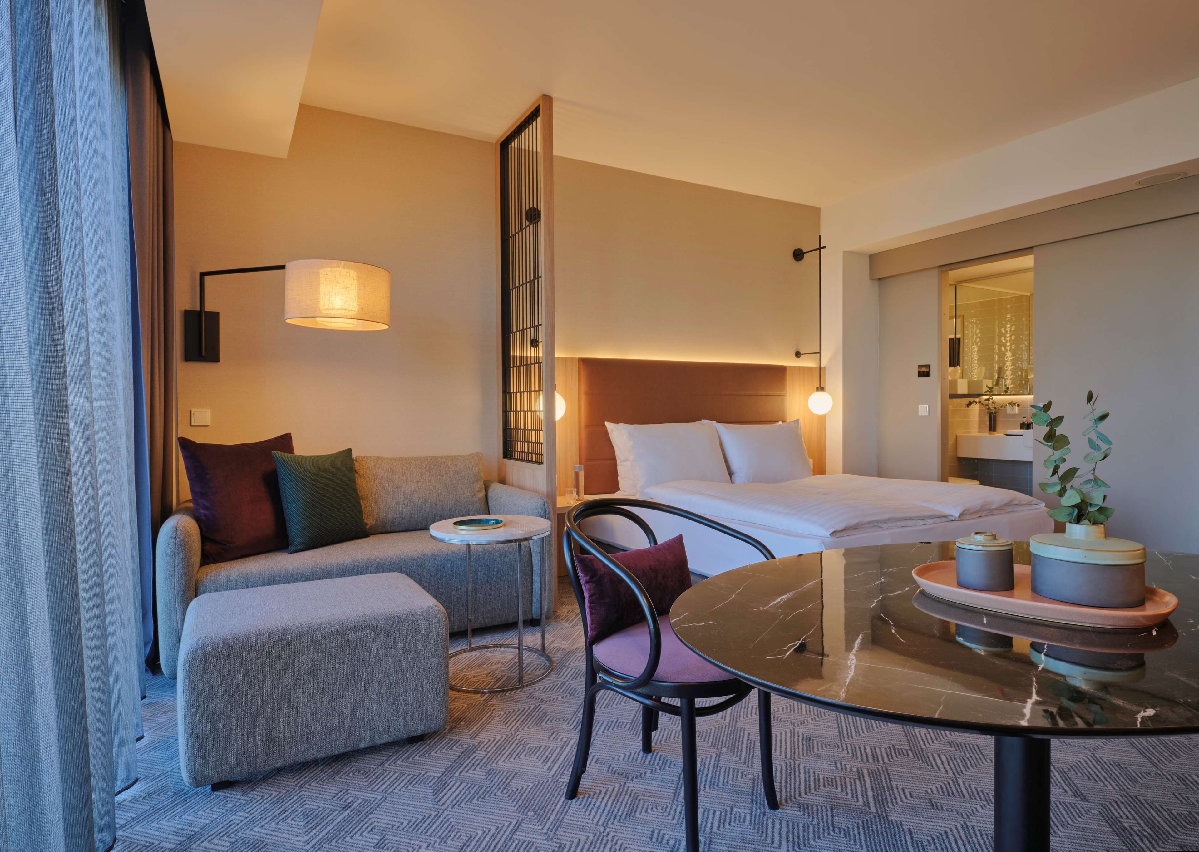 A hotel room with a bed, a sofa, a small table with two containers, a chair, and a lamp.