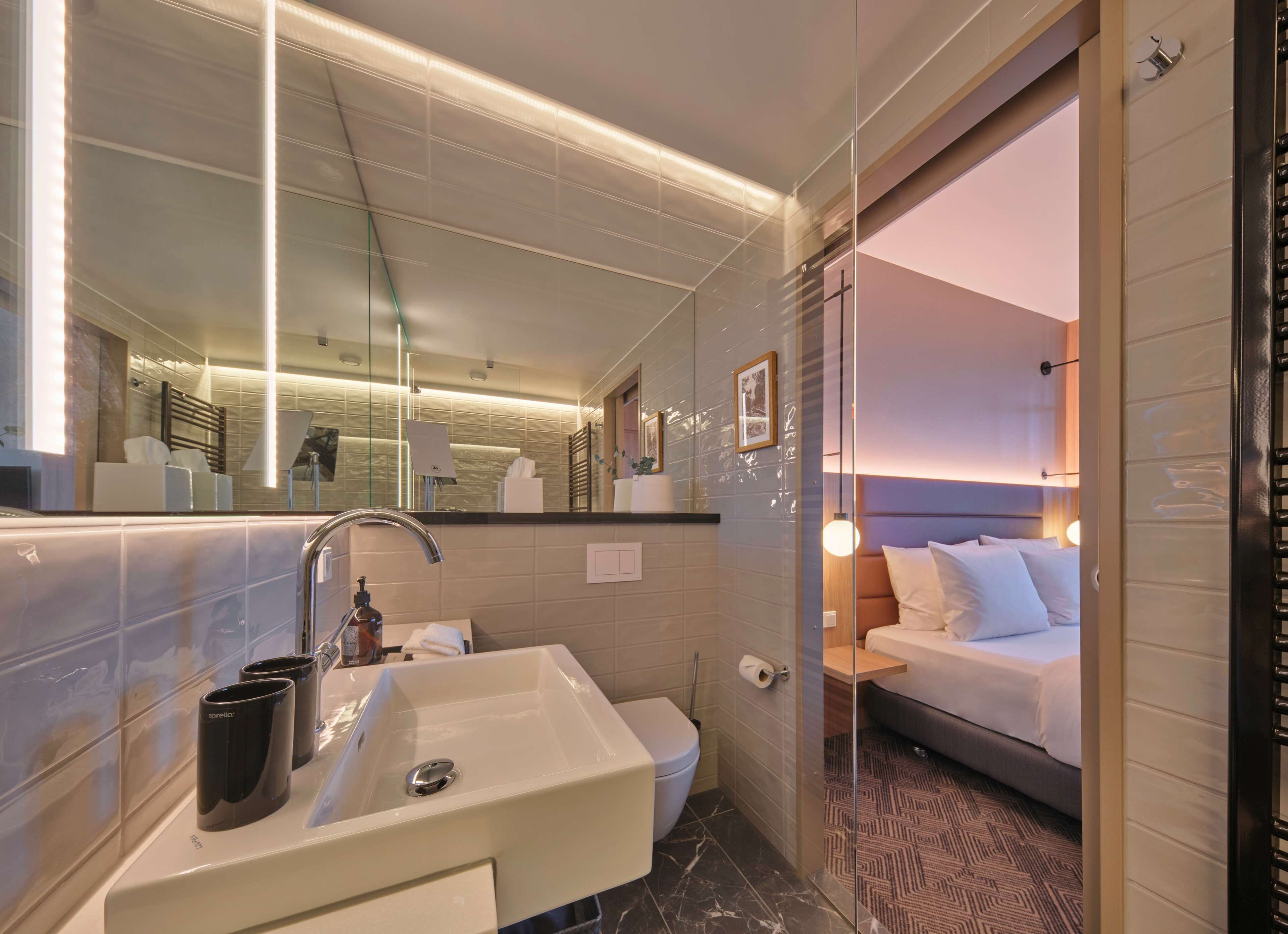Une salle de bain d’hôtel moderne, dotée d’un grand miroir, d’un lavabo et de toilettes. On peut apercevoir un lit à travers une porte en verre.