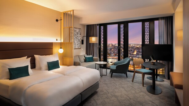 A hotel room with a bed, a sofa, a TV, and a view of the cityscape.