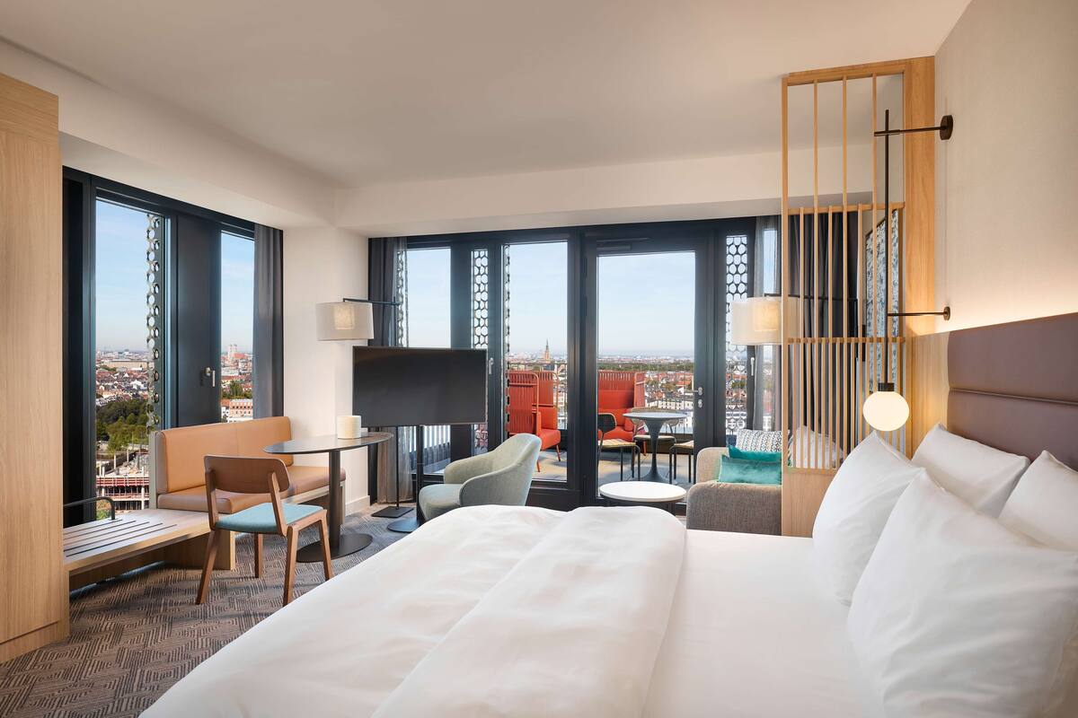 Ein Hotelzimmer mit einem großen Bett, einem Schreibtisch, einem Sessel, einer Couch und einem Fernseher. Das Zimmer hat große Fenster mit Blick auf die Stadt.