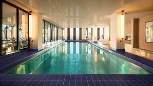 An indoor swimming pool with clear blue water, surrounded by walls with large windows and modern lighting fixtures.
