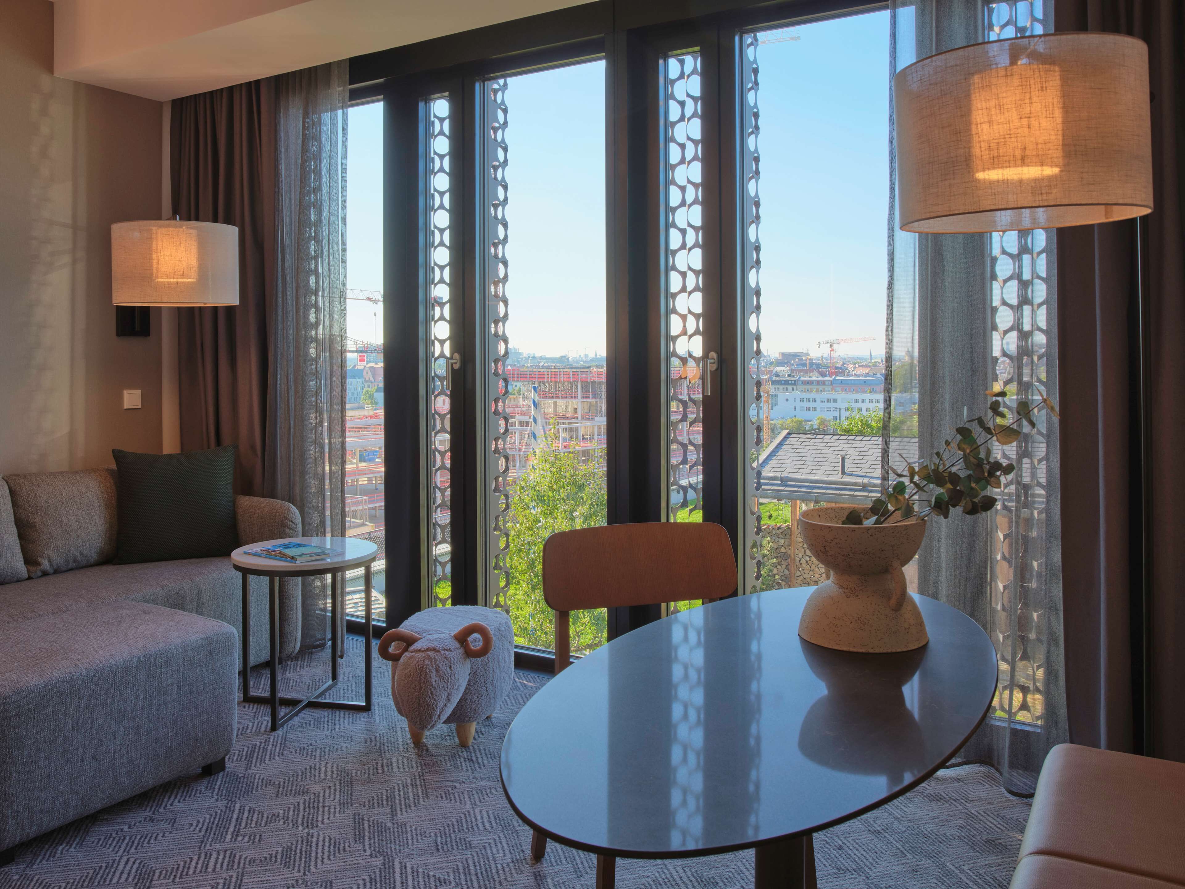 Ein modernes Hotelzimmer mit einer Couch, einem kleinen Tisch, einer dekorativen Schafskulptur und Blick auf die Stadt durch große Fenster.