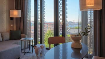 Ein modernes Hotelzimmer mit einer Couch, einem kleinen Tisch, einer dekorativen Schafskulptur und Blick auf die Stadt durch große Fenster.