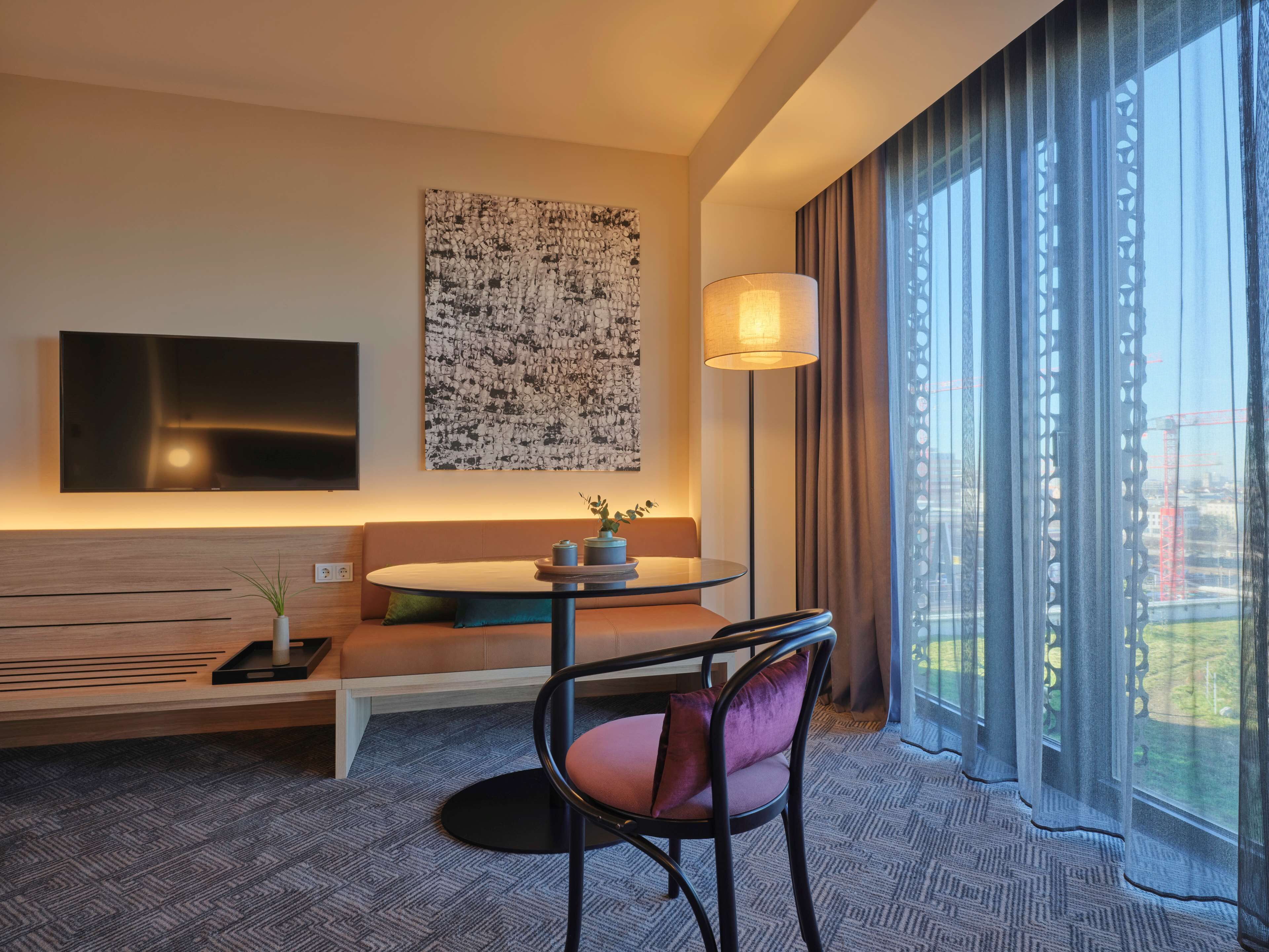 Et moderne hotelværelse med et fladskærms-TV monteret på væggen, et rundt bord med en vase og en lampe, en sofa med puder, og et stort vindue med udsigt over byen.
