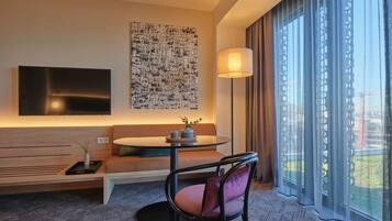 Et moderne hotelværelse med et fladskærms-TV monteret på væggen, et rundt bord med en vase og en lampe, en sofa med puder, og et stort vindue med udsigt over byen.