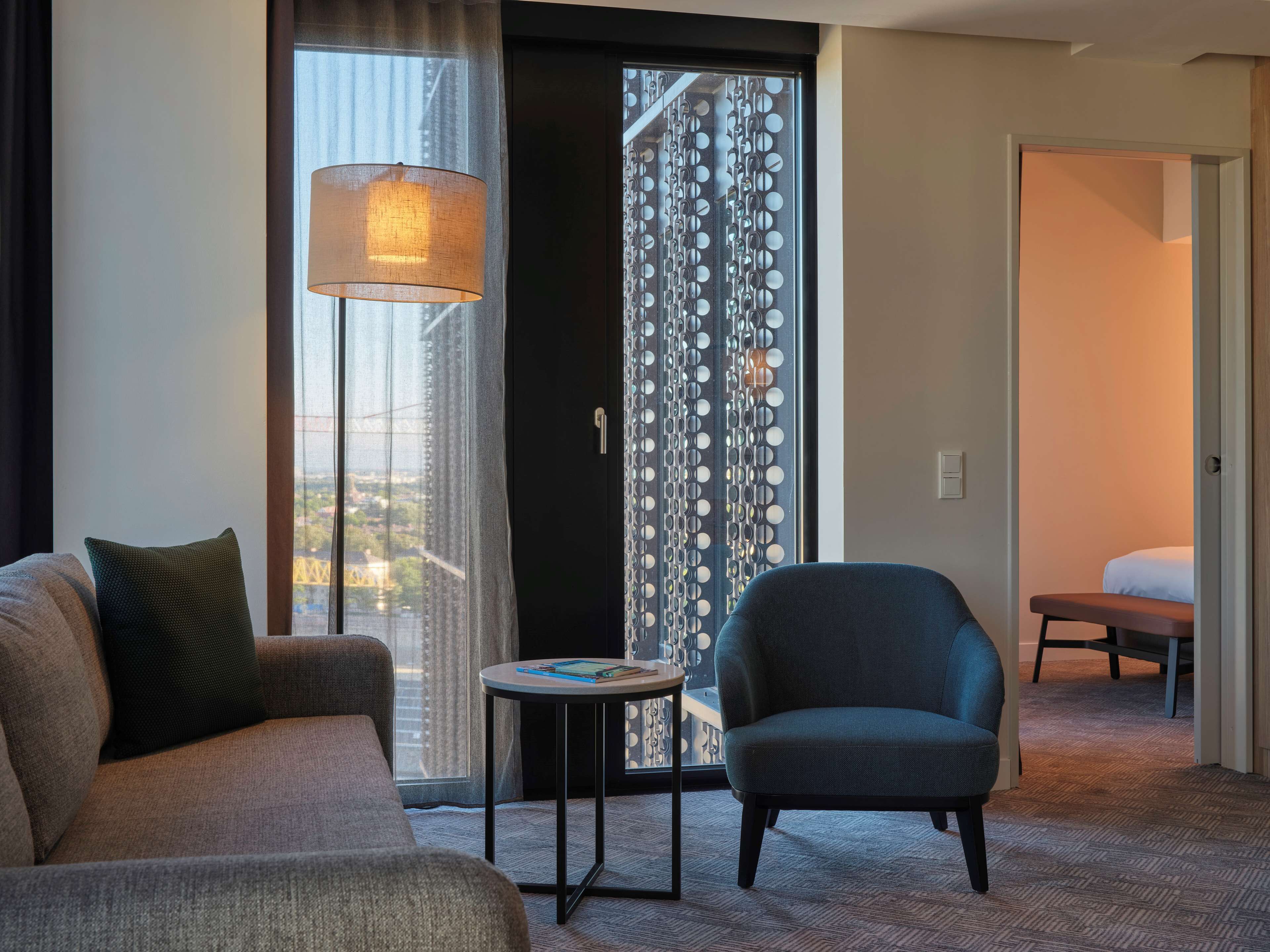 A hotel room with a sofa, armchair, small table, and a view of a cityscape through a large window.