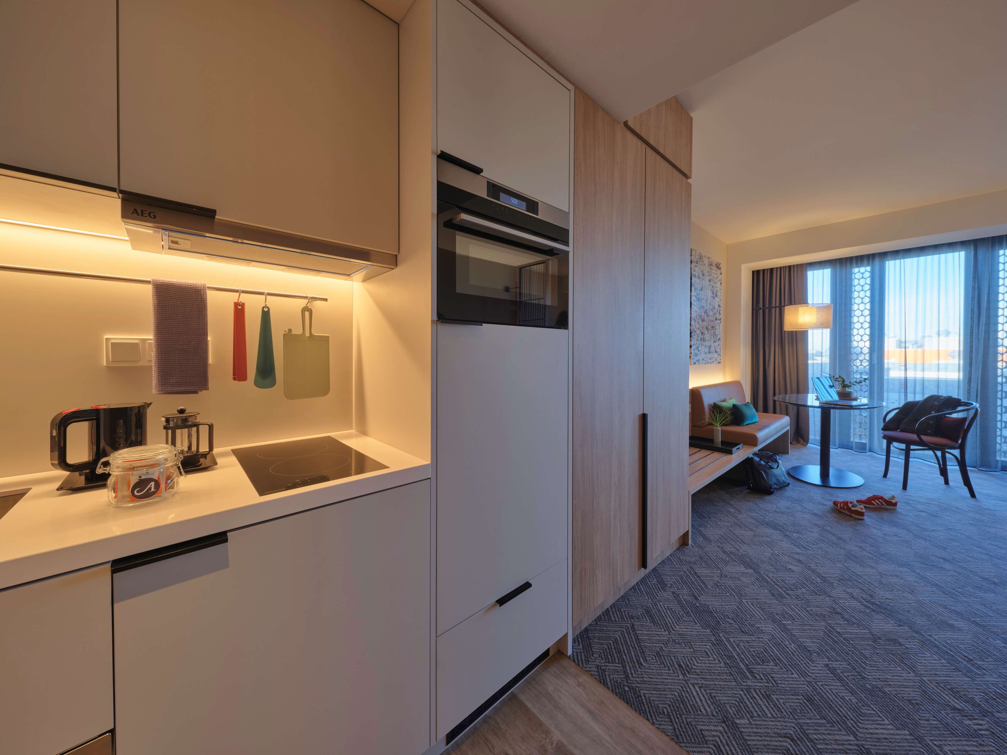 A modern hotel room with a kitchenette, a seating area with a table and chairs, and a large window with a view.