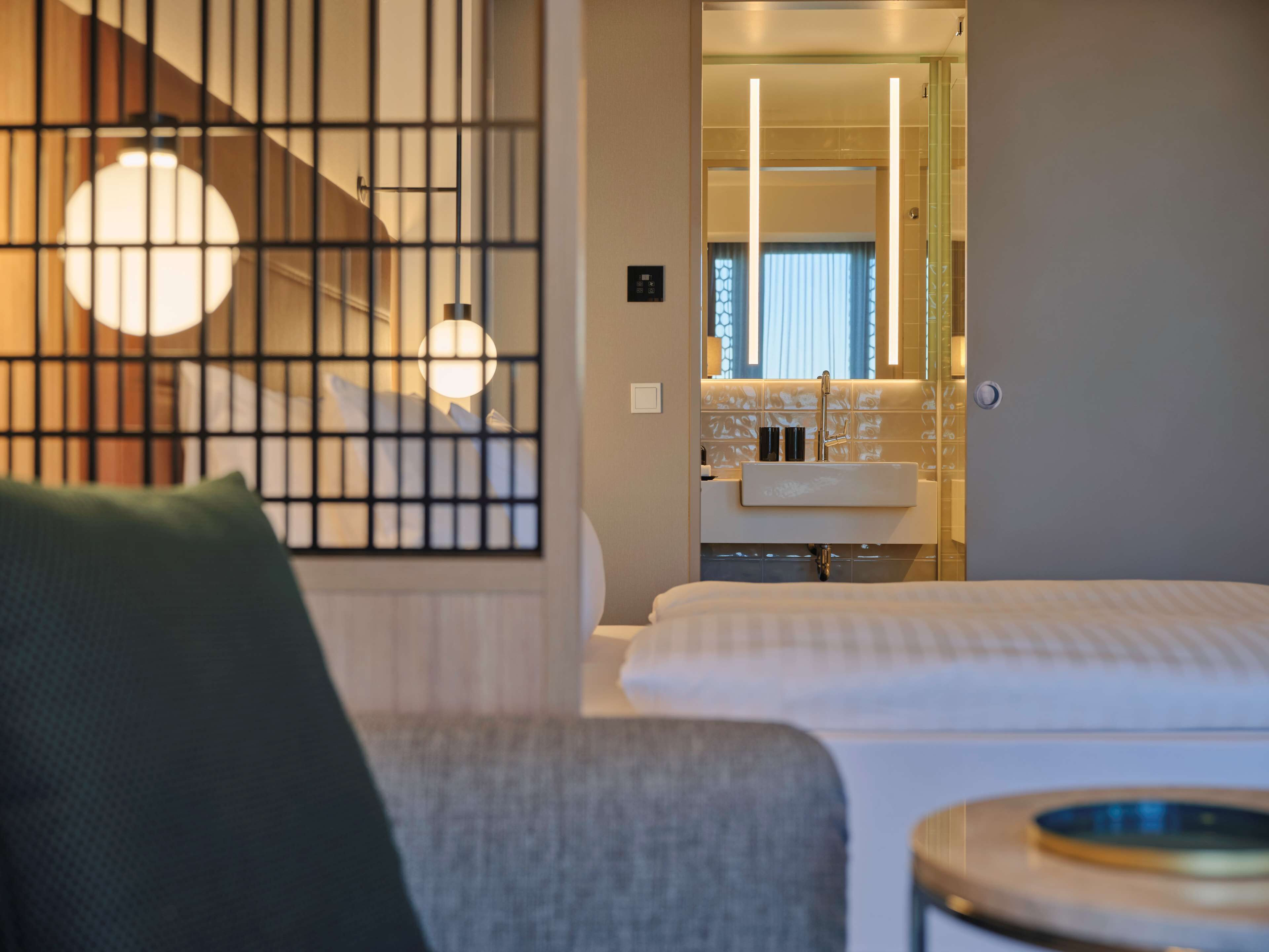 A hotel room with a bed, a chair, a small round table, and a view of a bathroom with a sink and mirror.