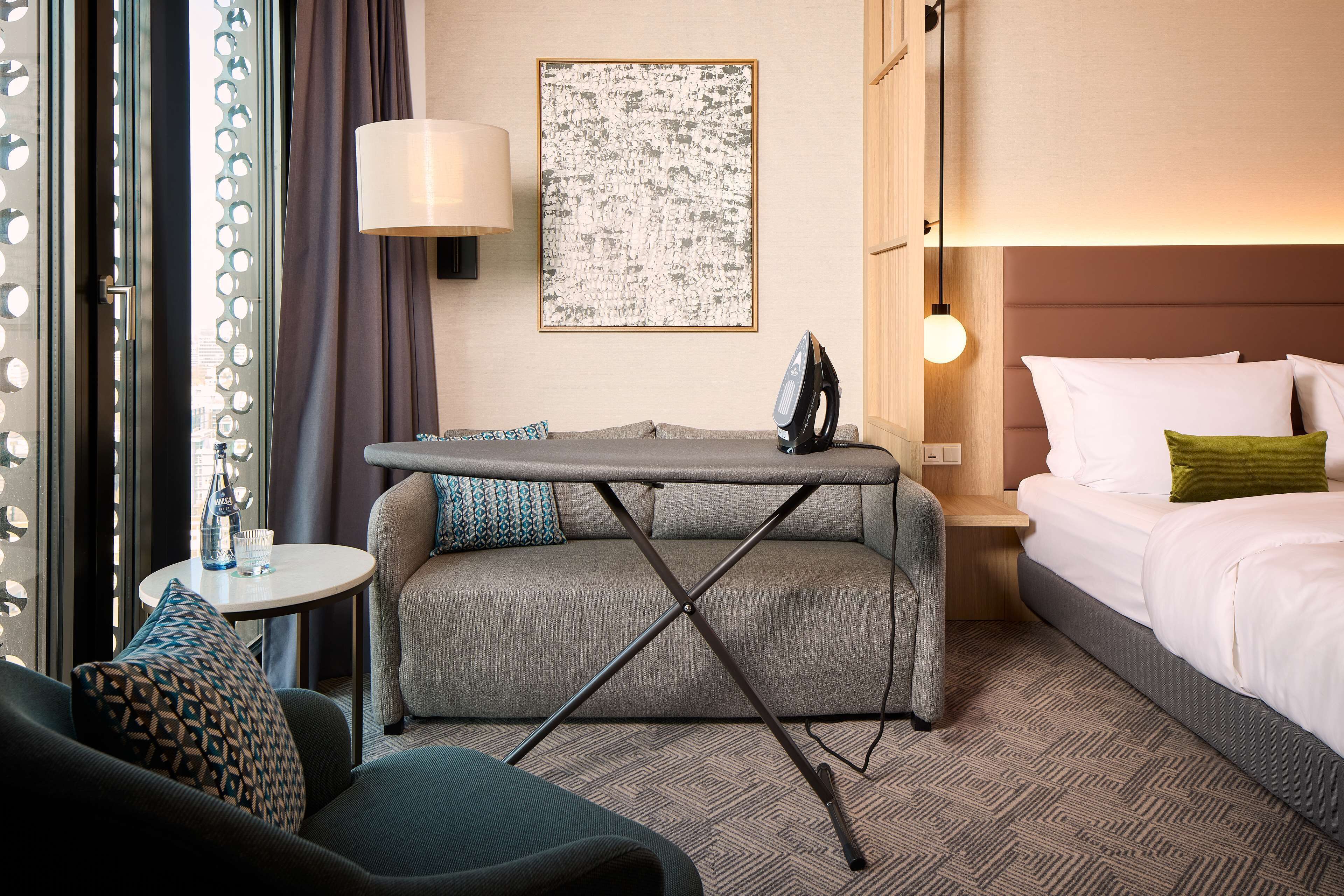 Una habitación de hotel con sofá, una mesita con lámpara, tabla de planchar, cama con almohadas y una obra de arte en la pared.