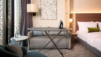 A hotel room with a sofa, a small table with a lamp, an ironing board, a bed with pillows, and a wall art piece.