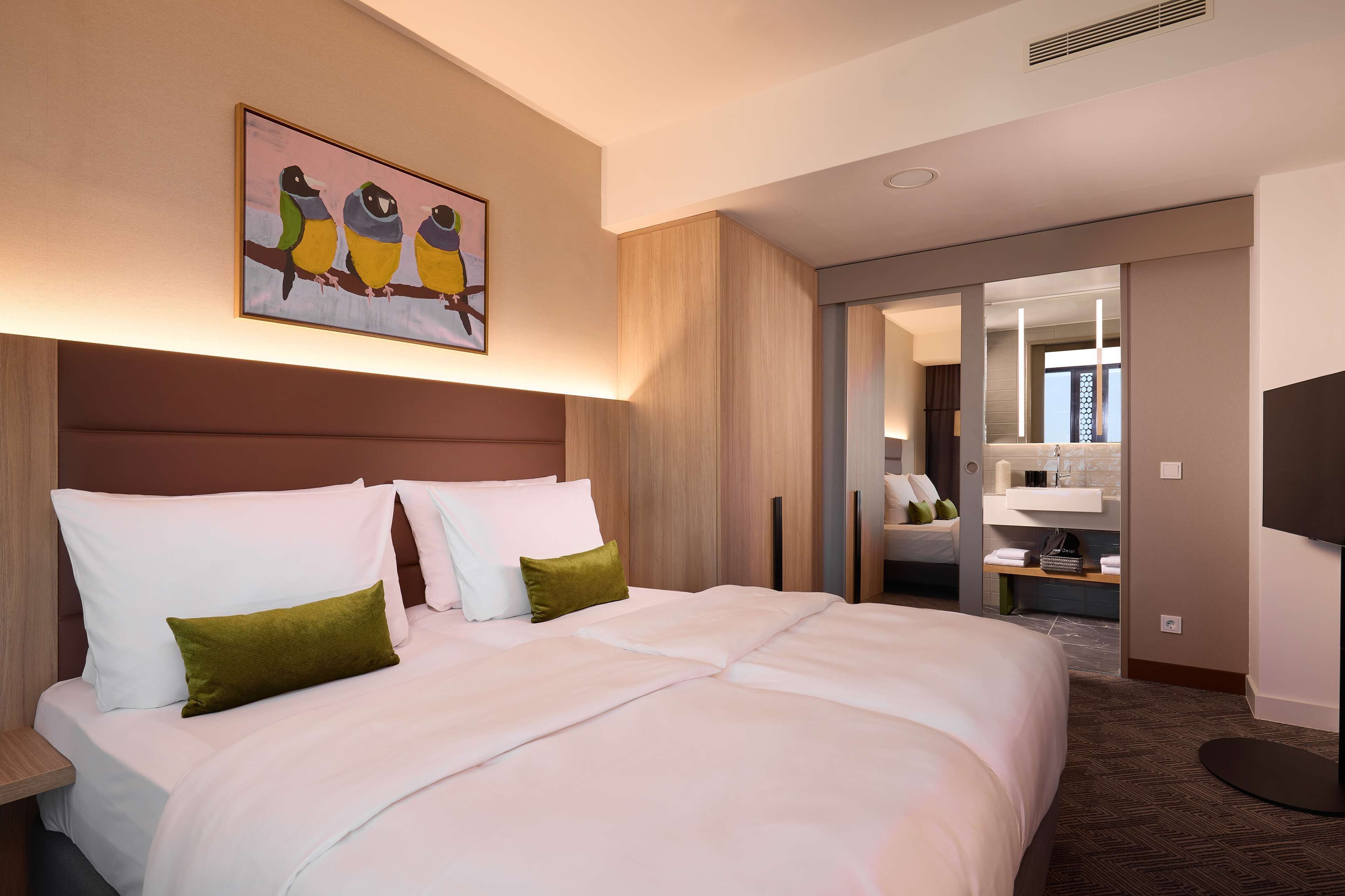 Quarto de hotel com uma cama grande, um quadro de pássaros na parede, um banheiro com espelho e uma televisão.
