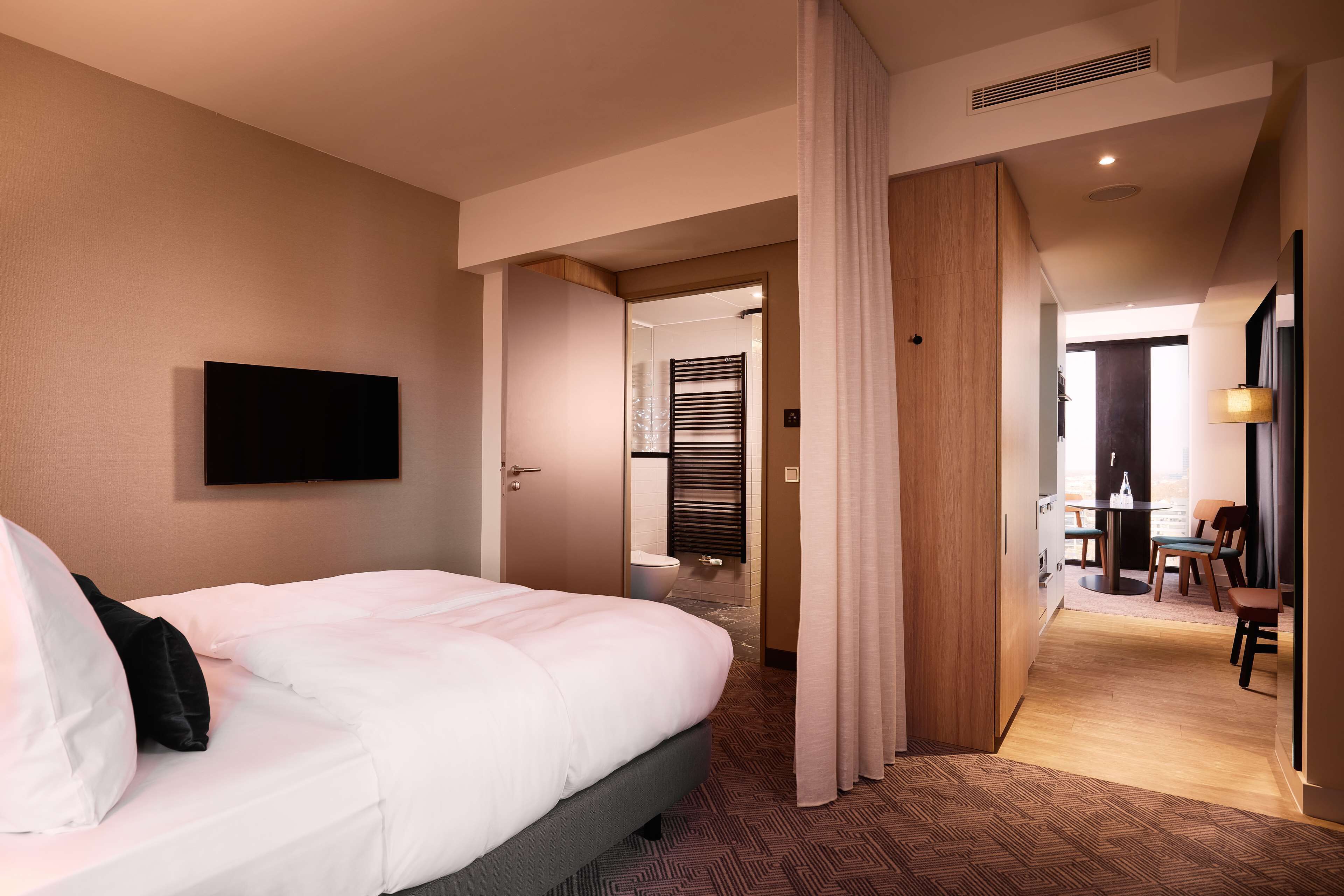Habitación de hotel con cama, televisor montado en la pared, puerta de madera y vista hacia un comedor.