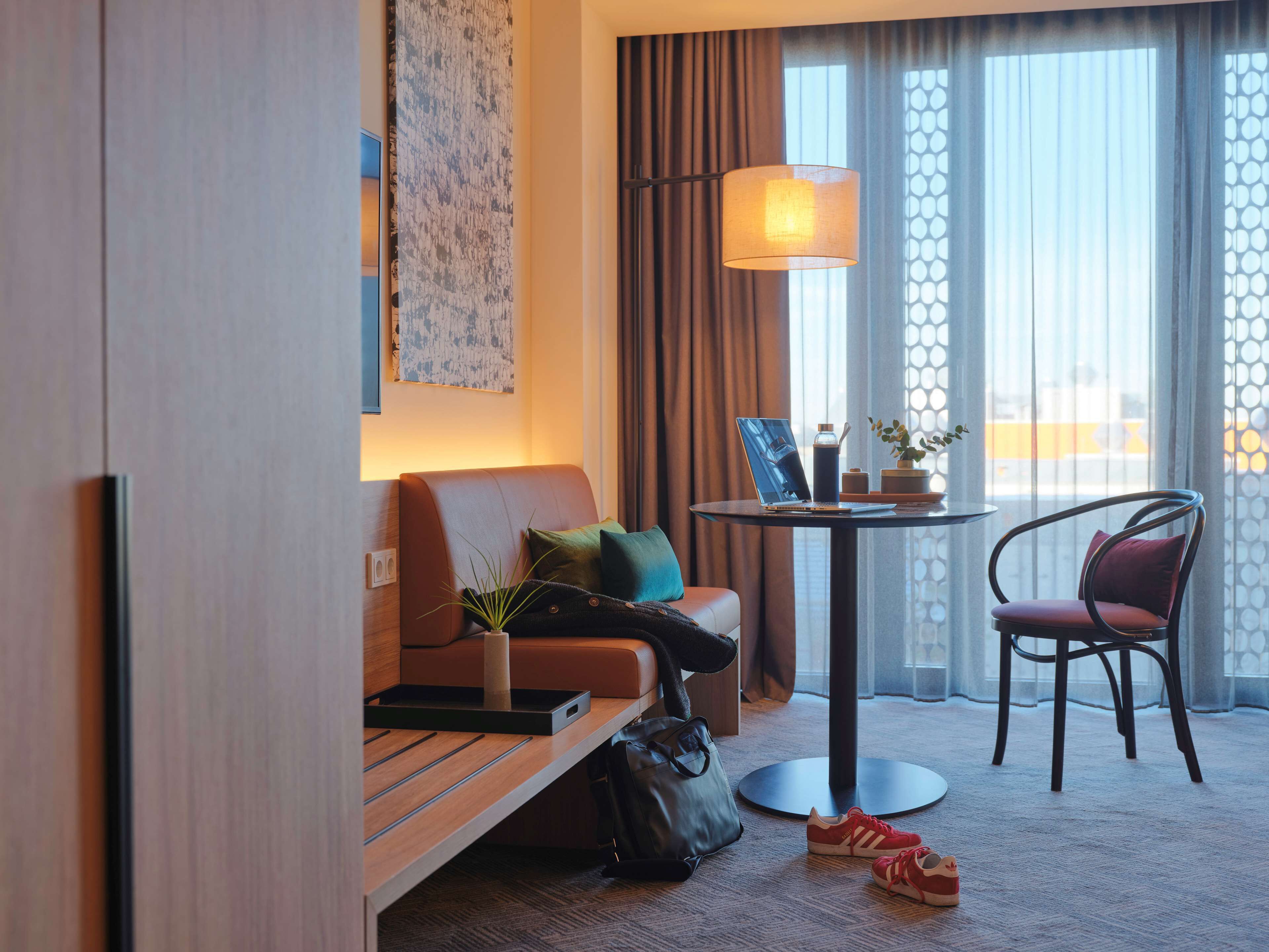 Een hotelkamer met een bank, een stoel, een klein tafeltje met een lamp, een laptop en een plant.