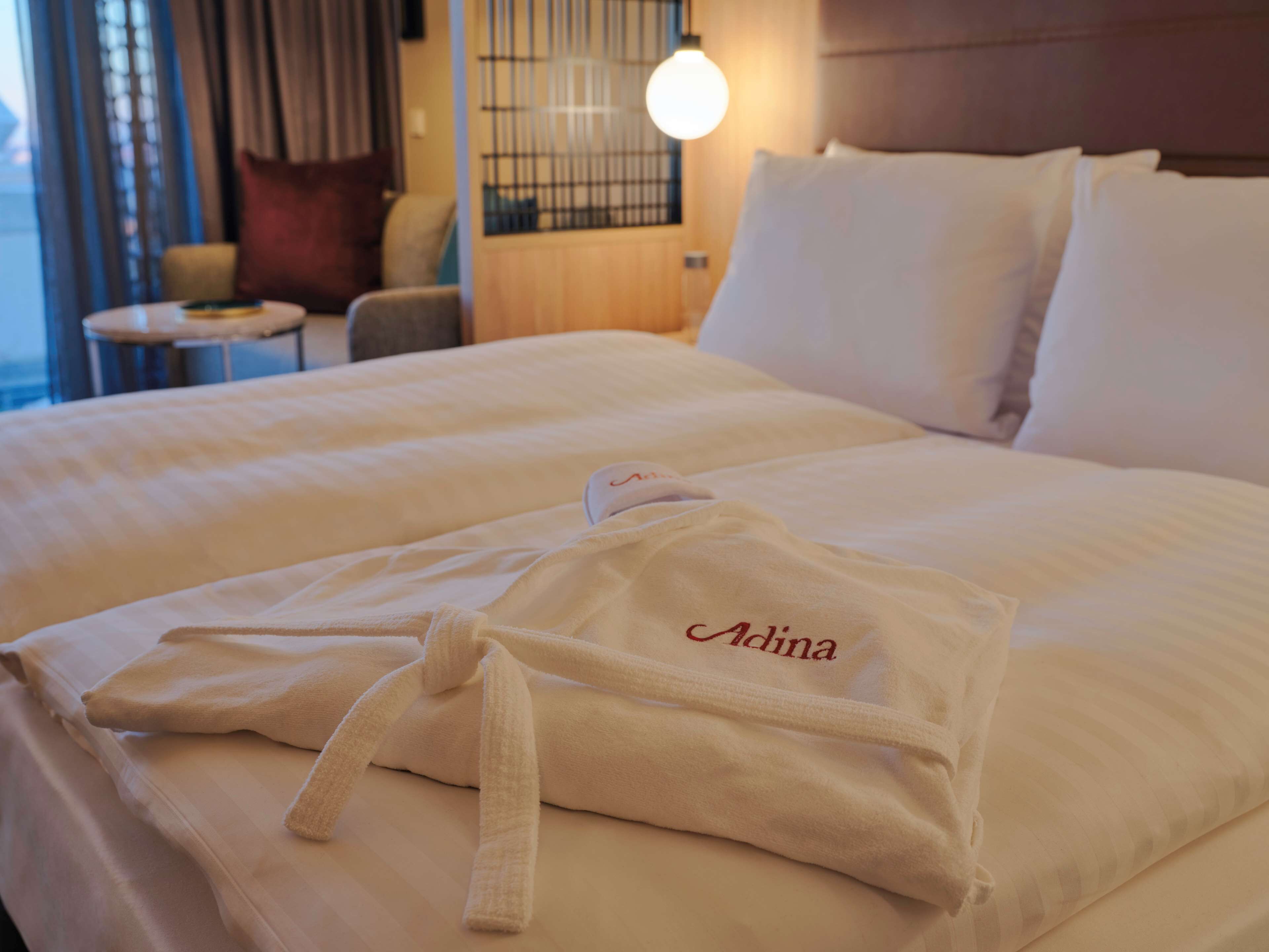 A hotel room with a neatly made bed, a robe with the name 'Adina' on it, and a lamp on the nightstand.
