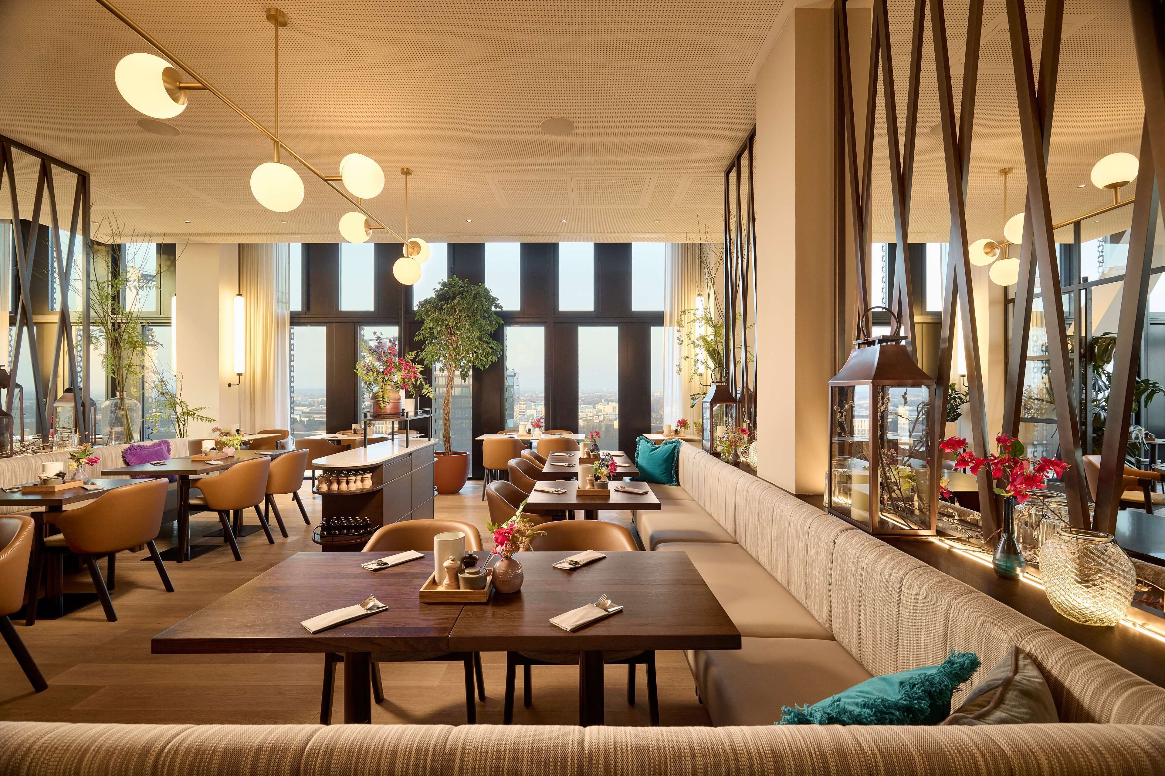 A modern restaurant with high ceilings, large windows, and a variety of seating options including tables and a long bench.