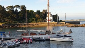 En lysthavn med flere både i havnen, en hvid bygning med rødt tag, og en klippefyldt kystlinje med træer i baggrunden.