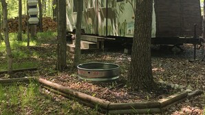 Un'area boscosa con un camper, un abbeveratoio in metallo e una pila di legna da ardere.