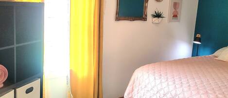 Un dormitorio con cama, cómoda, ventana con cortinas, espejo adosado a la pared y una planta pequeña.